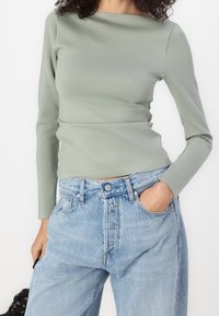 Persona che indossa un top a maniche lunghe verde salvia infilato in un paio di jeans di denim azzurri larghi, con una mano in tasca, su sfondo bianco.