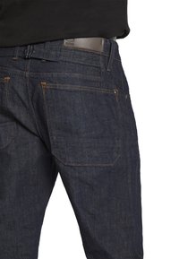 Vaqueros de mezclilla oscura con corte slim, costuras en contraste, bolsillos traseros y una etiqueta de cuero en la cinturilla con el logo de la marca.