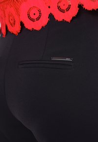 Pantalones negros con una tela negra texturizada, con un sobrecapa de encaje floral rojo en la cintura y un pequeño acento plateado en el bolsillo trasero.