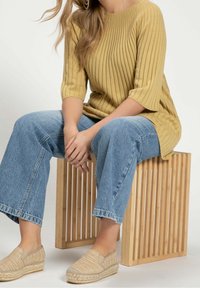 Gelbes geripptes Strickoberteil, hellblaue weite Jeans und beige Espadrilles. Model sitzt auf einem hölzernen Lattenstuhl.