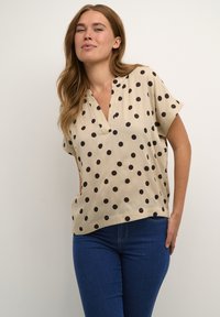Blouse beige à manches courtes avec des pois noirs, présentant un design en V et une coupe ample, fabriquée dans un tissu léger et lisse.