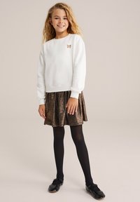 Witte sweatshirt met een klein gouden strikembleem, gecombineerd met een glanzende, bronzen geplooide rok, zwarte maillots en zwarte schoenen.