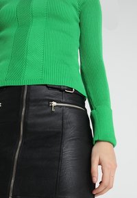 Grön ribbad stickad tröja kombinerad med en svart läderkjol med silverdragkedjor och midjebälte. Texturerad och figursydd design.