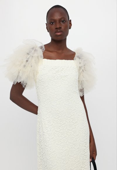 Witte kanten jurk met een vierkante halslijn en volumineuze lichtbeige tule pofmouwen, gedragen met een zwarte tas.