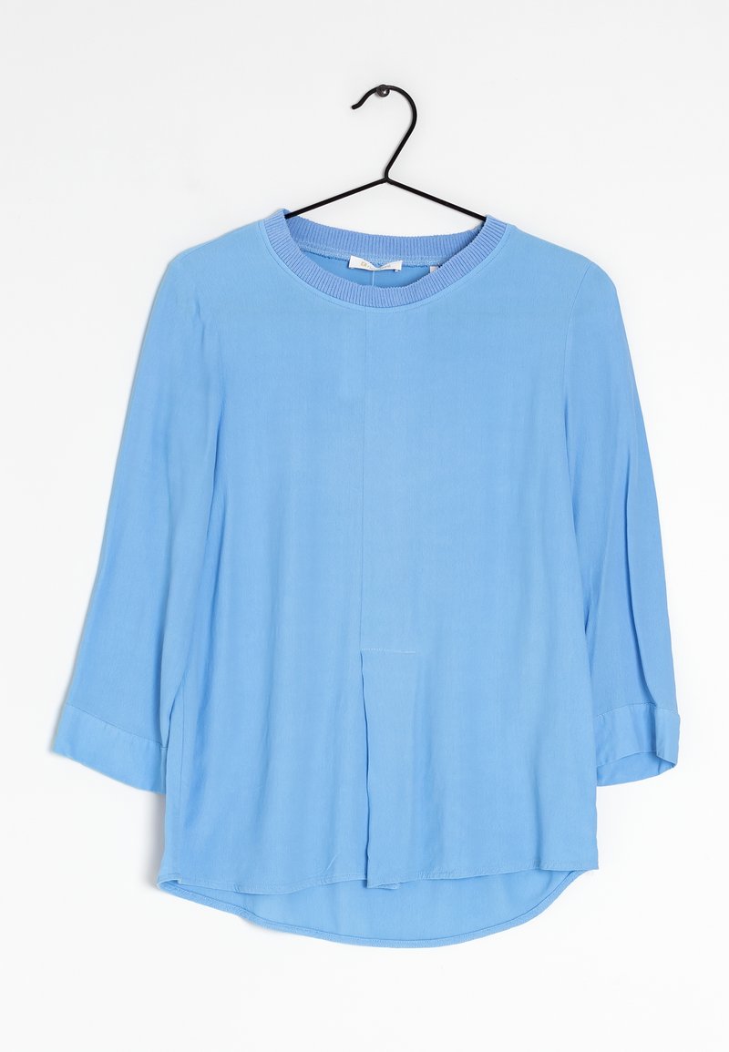 Himmelblaue Bluse mit langen Ärmeln und rundem Halsausschnitt, ausgestattet mit einer vorderen Naht und Faltendetails. Hergestellt aus einem glatten, weichen Stoff.