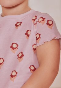 Hellrosa Kurzarmhemd mit einem wiederholten Blumenmuster in roten und pfirsichfarbenen Tönen, einem leicht gewellten Saum und geripptem Ausschnitt.