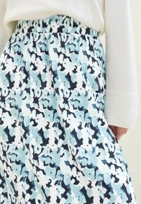 Nahaufnahme eines plissierten Rocks mit blauem, weißem und schwarzem abstraktem Blumenmuster, getragen mit einem weißen Pullover.