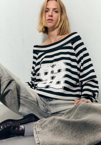 Młoda kobieta z blond włosami do ramion siedzi oparta o ścianę, ubrana w czarno-białą poszarpaną bluzkę bez ramiączek z nadrukiem "92" i jasnoszare dżinsy.