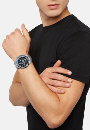 Sølv chronograph ur med sort urskive og blå accenter, udstyret med en armbånd i rustfrit stål. Båret på et håndled med lys hudtone.