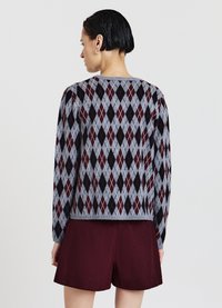 Gestrickter Pullover mit einem grauen, schwarzen und burgunderroten Rautenmuster. Mit langen Ärmeln und rundem Ausschnitt. Kombiniert mit bordeauxroten Shorts.