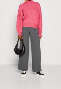 Personne portant un pull col roulé rose, un pantalon large gris, des baskets noir et blanc, tenant un petit sac à main noir à ses côtés.