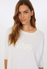 White short-sleeve sweater featuring a textured "PARIS" design in raised white letters, round neckline, and soft knit material.