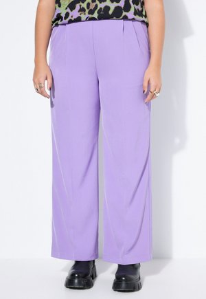 Person iført lyse lilla bukser med vide ben, sort chunky støvler og en grøn og sort leopardprint top, står op ad en hvid væg.