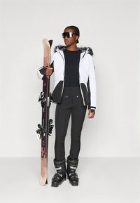Ski outfit featuring a white insulated jacket with a fur-lined hood, black pants, and ski boots. A ski held in one hand completes the look.