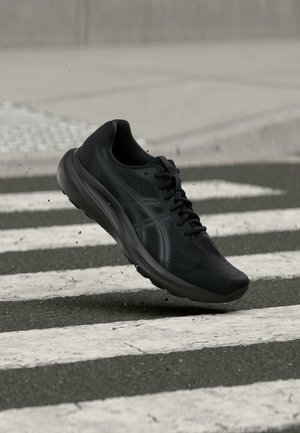 Chaussure de sport noire avec un design en mesh flottant au-dessus d’un passage piéton sur une rue asphaltée.
