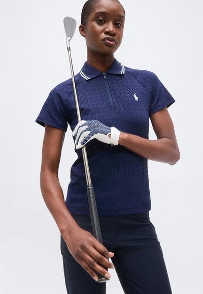 Γυναίκα που φορά μπλε σκούρο γκολφ πουκάμισο και γάντι, κρατώντας ένα γκολφ κλαμπ κάθετα μπροστά σε απλό φόντο.