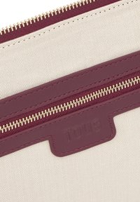 Cartera de tela beige con acentos de cuero burdeos, con cremalleras doradas y un logo "TOUS" en relieve. Diseño texturizado y moderno.