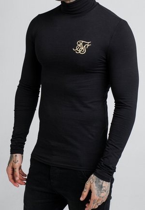 Uomo che indossa un dolcevita nero aderente con un piccolo logo bianco ricamato sul petto, con mani e braccia tatuate in vista.