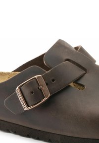 Brun lädersandal med ett brett rem och justerbar spänne, med en texturerad innersula och en gummisula. Varumärket syns på spännet.