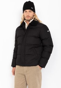 Sort quilted jakke med en blød sort krave, lange ærmer, lynlåslukning og to sidelommer, båret over en lys beige skjorte.