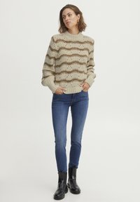 Beige Strickpullover mit wellenförmigen braunen Streifen, gerippten Bündchen und hohem Kragen. Kombiniert mit blauen Skinny-Jeans und schwarzen Knöchelstiefeln.