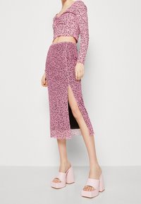 Žena oblečená v růžovém květovaném topu s dlouhým rukávem a odpovídající midi sukni se šikmým rozparkem, doplněná světle růžovými platformovými podpatky.