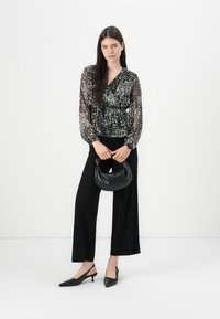 Blusa avvolgente nera in pizzo floreale abbinata a pantaloni neri a gamba larga. La modella tiene una piccola borsa nera e indossa tacchi neri a punta.