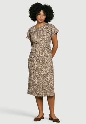 Frau mit lockigem Haar, die ein beige Midi-Kleid mit kurzen Ärmeln und Leopardenmuster sowie braune, niedrig absätzige Schuhe trägt und vor einfarbigem Hintergrund steht.