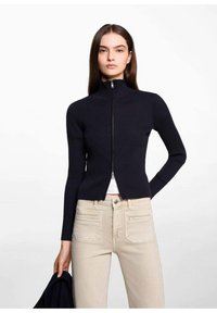 Mornarsko rebrasto zip-up pulover s visokim ovratnikom, prilegajočimi dolgimi rokavi in skrajšano zasnovo, v kombinaciji s svetlimi kavbojkami.