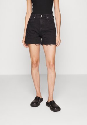 Persona che indossa pantaloncini di jeans neri a vita alta con orli sfrangiati, scarpe slip-on nere e una canotta nera senza maniche su sfondo semplice.
