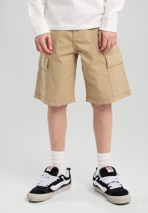 Beige cargo shorts dengan saku samping, potongan lurus, dan jahitan hem, dipadukan dengan sepatu sneaker hitam dan putih serta kaus kaki crew putih.