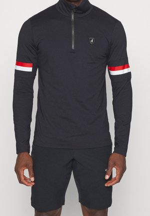 Uomo che indossa una maglia nera a maniche lunghe con zip a quarto e strisce rosse e bianche sulle maniche e pantaloncini neri, in piedi su uno sfondo semplice.