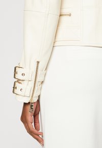 Veste en cuir crème avec quincaillerie dorée, dotée d'un poignet zippé et de sangles ajustables. Texture lisse et coupe cintrée.