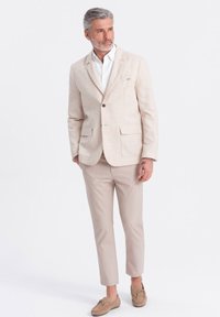 Blazer de lino beige claro con dos bolsillos delanteros, combinado con pantalones ajustados a juego. Camisa blanca debajo, complementada con mocasines marrones.