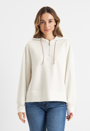 Crème-kleurige zip-up hoodie met een frontale rits, verstelbare roze trekkers en lange mouwen. Gecombineerd met blauwe spijkerbroek.