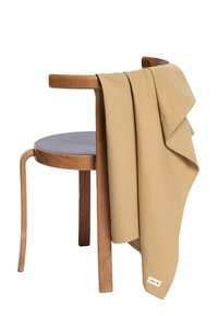 Texturovaný hořčicově žlutý pléd přehozený přes dřevěnou židli s zahnutými nohami a kulatým sedákem, s tkaným vzorem a detaily s visačkou.