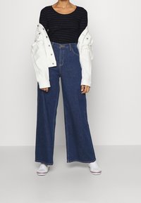 Černý pruhovaný top s dlouhým rukávem, bílá oversize bunda, džíny s vysokým pasem a širokými nohavicemi tmavě modré barvy a bílé tenisky s červenými akcenty.