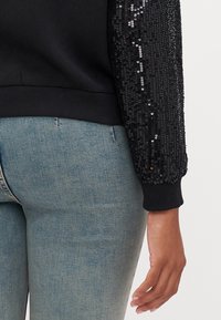 Czarna bluza z cekinowymi rękawami, ściągaczami na mankietach i u dołu. Noszona z jasnoniebieskimi dopasowanymi dżinsami.