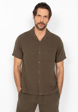 Mand med skæg iført en brun kortærmet skjorte med knapper og matchende bukser, stående med hænderne i lommerne mod en hvid baggrund.