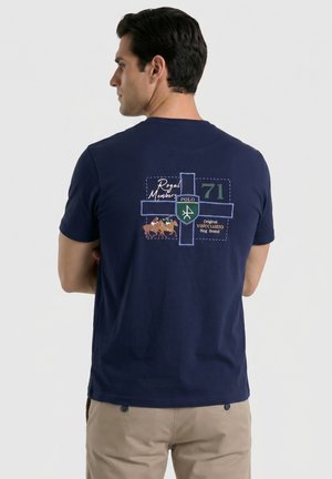 Hombre de espaldas, vistiendo una camiseta azul marino con gráfico de caballo de polo, escudo verde y blanco, y texto impreso en la espalda.
