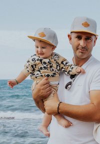 Siva snapback kapa z rumenimi akcenti; bež obleka z vzorcem pand. Odrasla oseba drži otroka ob morju, z vodo v ozadju.