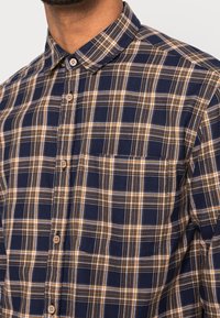 Camisa de cuadros azul y beige en tejido de algodón. Presenta un collar con botones, bolsillo en el pecho y cierres de botones con detalles de madera.