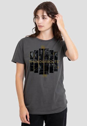 Woman wearing dark gray Thunderbolts graphic t-shirt, touching hair, looking slightly to the side against a plain light background.