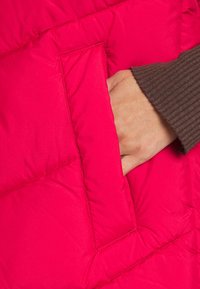 Heldere roze pufferjack met een diagonale quiltpatroon en een zijzak, met een bruine geribbelde mouw zichtbaar nabij de zak.