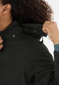 Zwarte waterdichte jas met hoge kraag, grip sluiting en verstelbaar trekkoord. Bevat knoppen en een gladde, matte textuur.