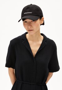 Schwarze Kappe mit dem weißen Stickmotiv "PERMANÈNT", getragen zu einem strukturierten schwarzen Hemdblusenkleid mit kurzen Ärmeln und einem taillierten Gürtel.
