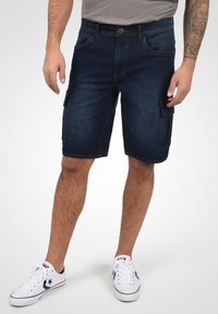 Bermudas de mezclilla, azul oscuro, de longitud media con bolsillos laterales, pasadores y detalles cosidos. Combinadas con zapatillas blancas.