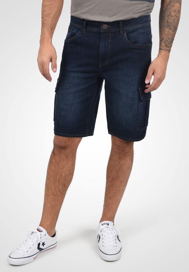 Bermudas de mezclilla, azul oscuro, de longitud media con bolsillos laterales, pasadores y detalles cosidos. Combinadas con zapatillas blancas.