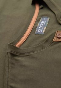 Camiseta verde oliva con un ribete de color marrón en el cuello, que presenta una etiqueta con la marca "ombre". La tela es suave y de textura lisa.