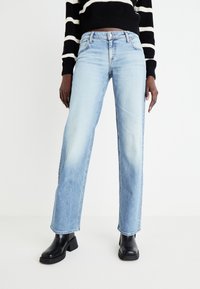 Light blue straight-leg jeans made of denim. Features a faded wash, five pockets, and a button closure. Worn with a black striped sweater.